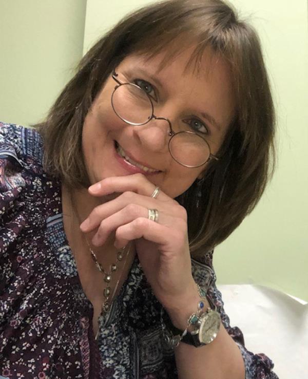 Jaye Murray seen in selfie, smiling at camera wearing round glasses, jewelry and printed shirt.