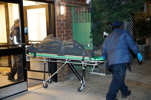 Two ambulance workers wheeling a stretcher with a black body bag on it outside a building.