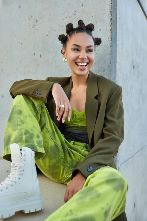 Positive carefree teenage girl with bun hairstyle looks happily away wears stylish green costume and white boots blue eyeliner feels good poses against grey co<em></em>ncrete wall. Urban lifestyle concept