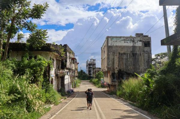 A street in Marawi. It is lined by ruined and abando<em></em>ned buildings. Plants are growing out of them. Two people are walking down the middle of the street away from the camera. There are no cars and noone else is around.