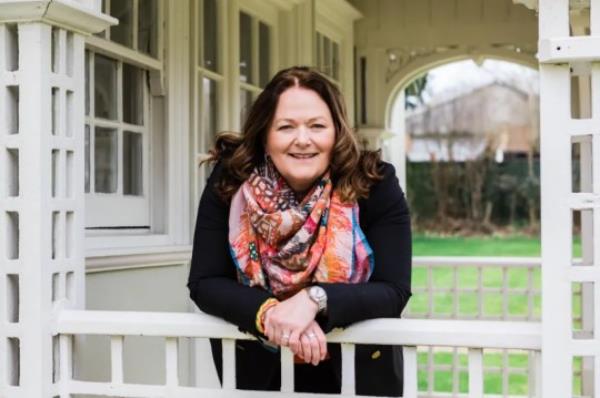 Alison is leaning on a garden porch fence, looking to camera and smiling.