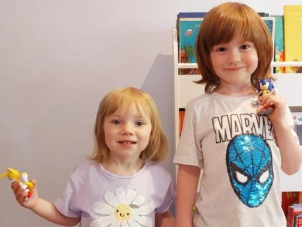 Sarah's kids are standing next to each other, wearing white t-shirts, looking to camera and holding a toy in each hand