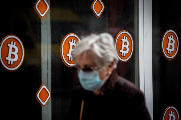 Elderly pedestrian wearing a face mask walking past a Bitcoin ATM kiosk in Barcelona, Spain.