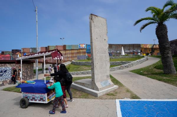 A family pushes a snack cart past a slab of the Berlin Wall, displayed near the border wall that separates the United States from Mexico, in Tijuana, Mexico, Friday, Aug. 25, 2023.