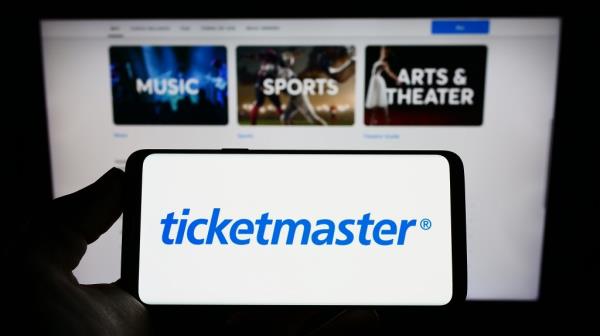 Person holding a cellphone displaying the logo of Ticketmaster Entertainment Inc, with a business webpage in the background.