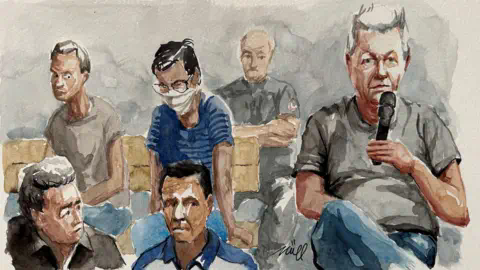 Reuters A court sketch depicting Dominique Pelicot on the right hand side, a man with grey hair, wearing a grey T-shirt and blue trousers, sitting up straight and holding a microphone. Behind him is a uniformed man standing guard and four other men, with uncertain ex<em></em>pressions of their faces or hanging their heads, who may be some of the co-defendants.