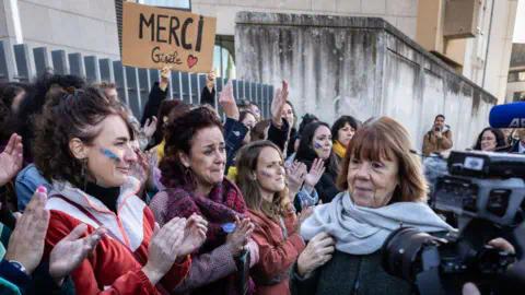 Getty Images Gisele Pelictor, a woman with auburn hair in a bob, wearing a coat and a pale blue scarf, surrounded by a crown of women on one side applauding her, some who seem overcome with emotion, and one person holding a sign saying, 